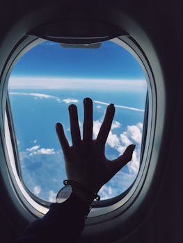 Silhouette of a hand touching an airplane window with a view of clouds and the sky.